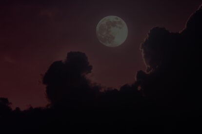 Full moon rising behind dark clouds at dusk with a crimson sky—moody night scene?