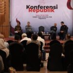 Speaker addresses audience from podium on a conference stage panelists sit in a row behind flags nearby banner reads konferensi republik in the background
