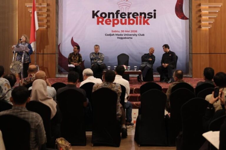 Speaker addresses audience from podium on a conference stage; panelists sit in a row behind, flags nearby, banner reads Konferensi Republik in the background.