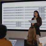 Woman presenter speaks into a microphone while holding papers standing in front of a large screen filled with spreadsheet style charts audience seated in foreground