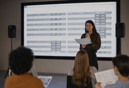 Woman presenter speaks into a microphone while holding papers, standing in front of a large screen filled with spreadsheet-style charts; audience seated in foreground.