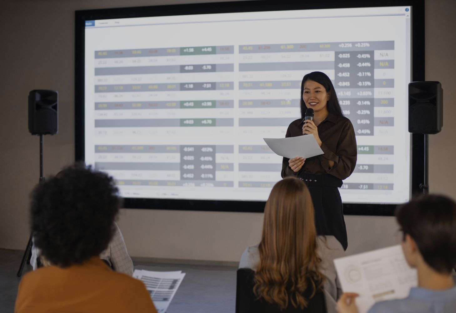 Woman presenter speaks into a microphone while holding papers standing in front of a large screen filled with spreadsheet style charts audience seated in foreground