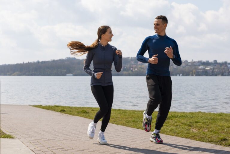 Two runners (a woman and a man) jogging along a brick path beside a lake on a sunny day with hills in the distance.