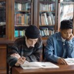 Two students study at a library table reading open books with tall wooden bookshelves behind them