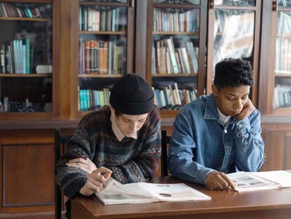 Two students study at a library table, reading open books with tall wooden bookshelves behind them.