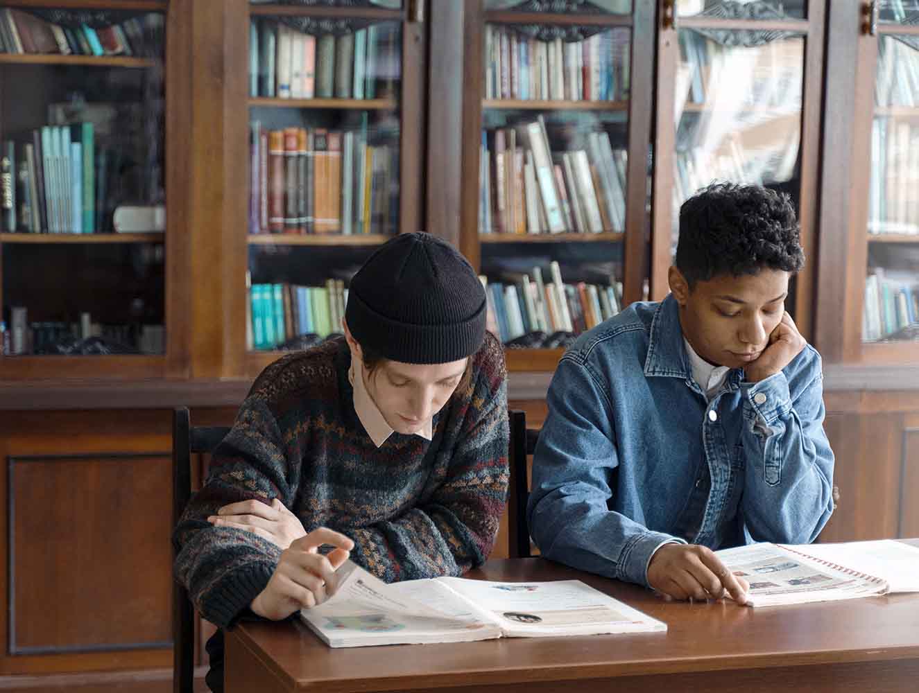 Two students study at a library table reading open books with tall wooden bookshelves behind them