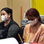 Two women wearing masks sit at a desk with papers and pencils in an indoor setting likely a classroom or office