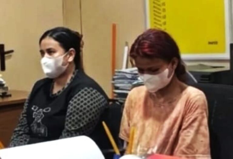 Two women wearing masks sit at a desk with papers and pencils in an indoor setting, likely a classroom or office.