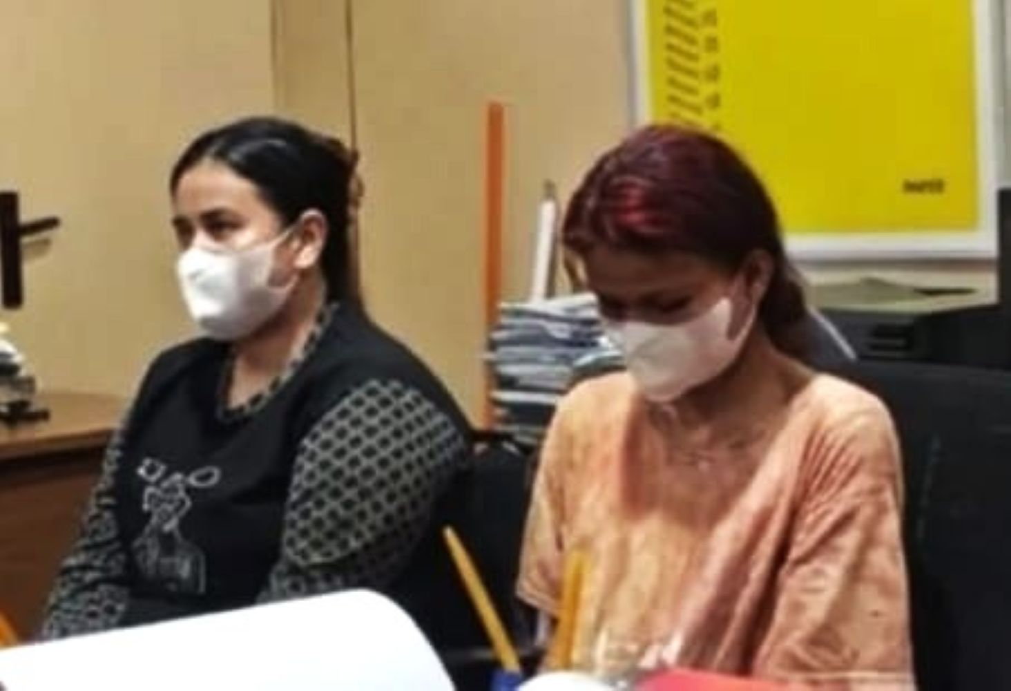 Two women wearing masks sit at a desk with papers and pencils in an indoor setting likely a classroom or office