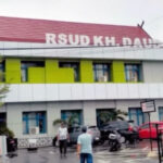Exterior of a colorful hospital building with a red roof and a visible rsud kh  sign with parked cars and a few people walking in the parking area