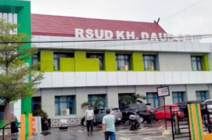 Exterior of a colorful hospital building with a red roof and a visible rsud kh  sign with parked cars and a few people walking in the parking area