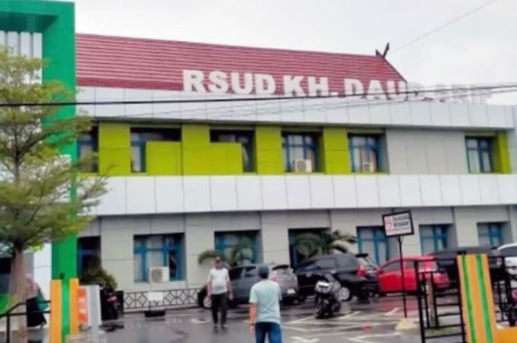 Exterior of a colorful hospital building with a red roof and a visible rsud kh  sign with parked cars and a few people walking in the parking area