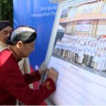 Man in red traditional clothing signs a large board showing a group photo at an outdoor event watched by onlookers behind him