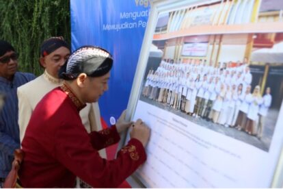 Man in red traditional clothing signs a large board showing a group photo at an outdoor event, watched by onlookers behind him.