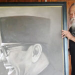Bearded elderly man with long white hair stands beside a large grayscale portrait of a smiling man wearing glasses and a top hat framing the image