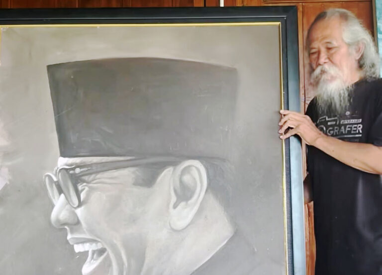 Bearded elderly man with long white hair stands beside a large grayscale portrait of a smiling man wearing glasses and a top hat framing the image.