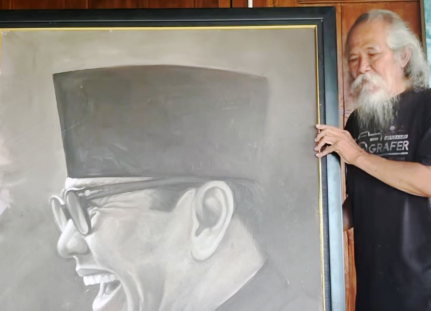 Bearded elderly man with long white hair stands beside a large grayscale portrait of a smiling man wearing glasses and a top hat framing the image