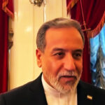 Mature man with gray hair and beard in a black suit at a formal event with red drapes in the ornate background