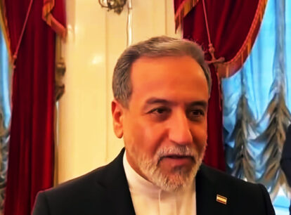 Mature man with gray hair and beard in a black suit at a formal event with red drapes in the ornate background