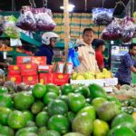 Busy market fruit stand with a pile of green avocados in the foreground and shoppers in the background
