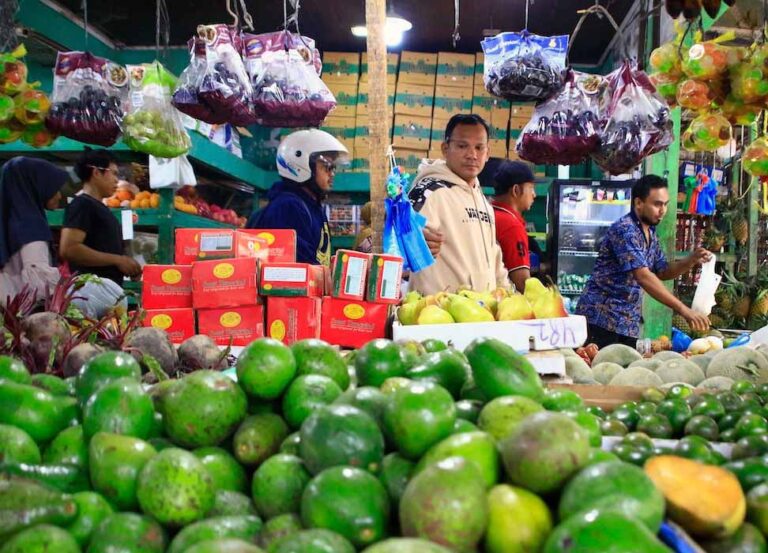 Busy market fruit stand with a pile of green avocados in the foreground and shoppers in the background.