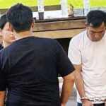 Man in a white tshirt is handcuffed standing with others outdoors near a wooden table with water bottles nearby