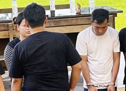 Man in a white tshirt is handcuffed standing with others outdoors near a wooden table with water bottles nearby