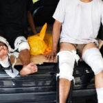 Two injured people sit in the open trunk of a car with leg casts and bandages on their legs one barefoot toward the edge of the bumper
