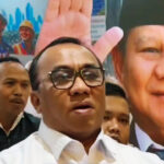 Central man wearing glasses and a white shirt speaks at a crowd with supporters and a large campaign poster behind him showing a smiling man and a raised hand A white heart icon is visible in the bottom right corner