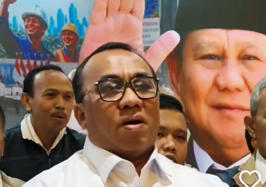 Central man wearing glasses and a white shirt speaks at a crowd with supporters and a large campaign poster behind him showing a smiling man and a raised hand A white heart icon is visible in the bottom right corner