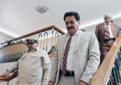 Three men in business attire descend a stairwell inside a building with the man in a light gray suit leading in front