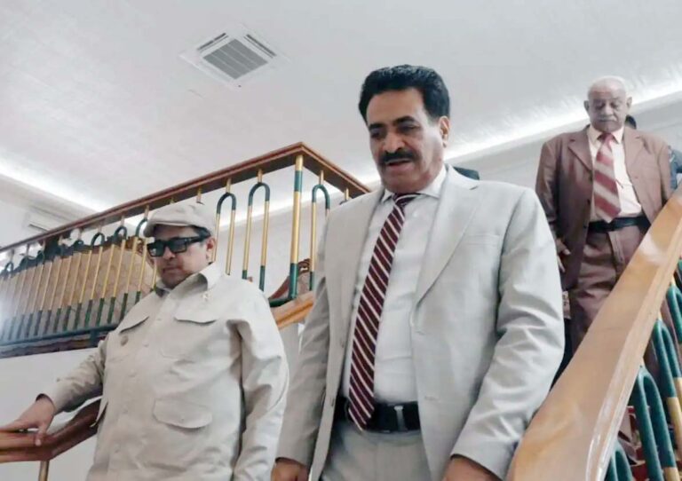 Three men in business attire descend a stairwell inside a building, with the man in a light gray suit leading in front.