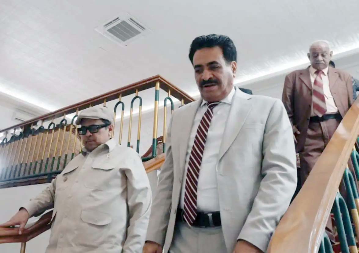 5ehaikal1 catrawarta Three men in business attire descend a stairwell inside a building with the man in a light gray suit leading in front