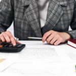 Person in a plaid blazer uses a calculator and writes on documents at a desk suggesting accounting work