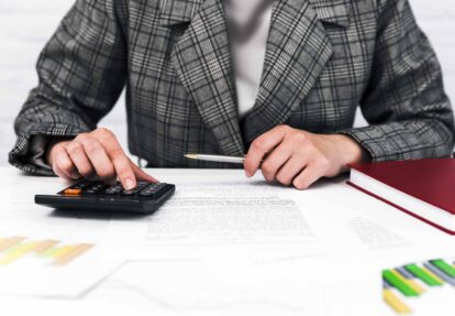 Person in a plaid blazer uses a calculator and writes on documents at a desk suggesting accounting work