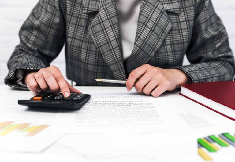 Person in a plaid blazer uses a calculator and writes on documents at a desk, suggesting accounting work.