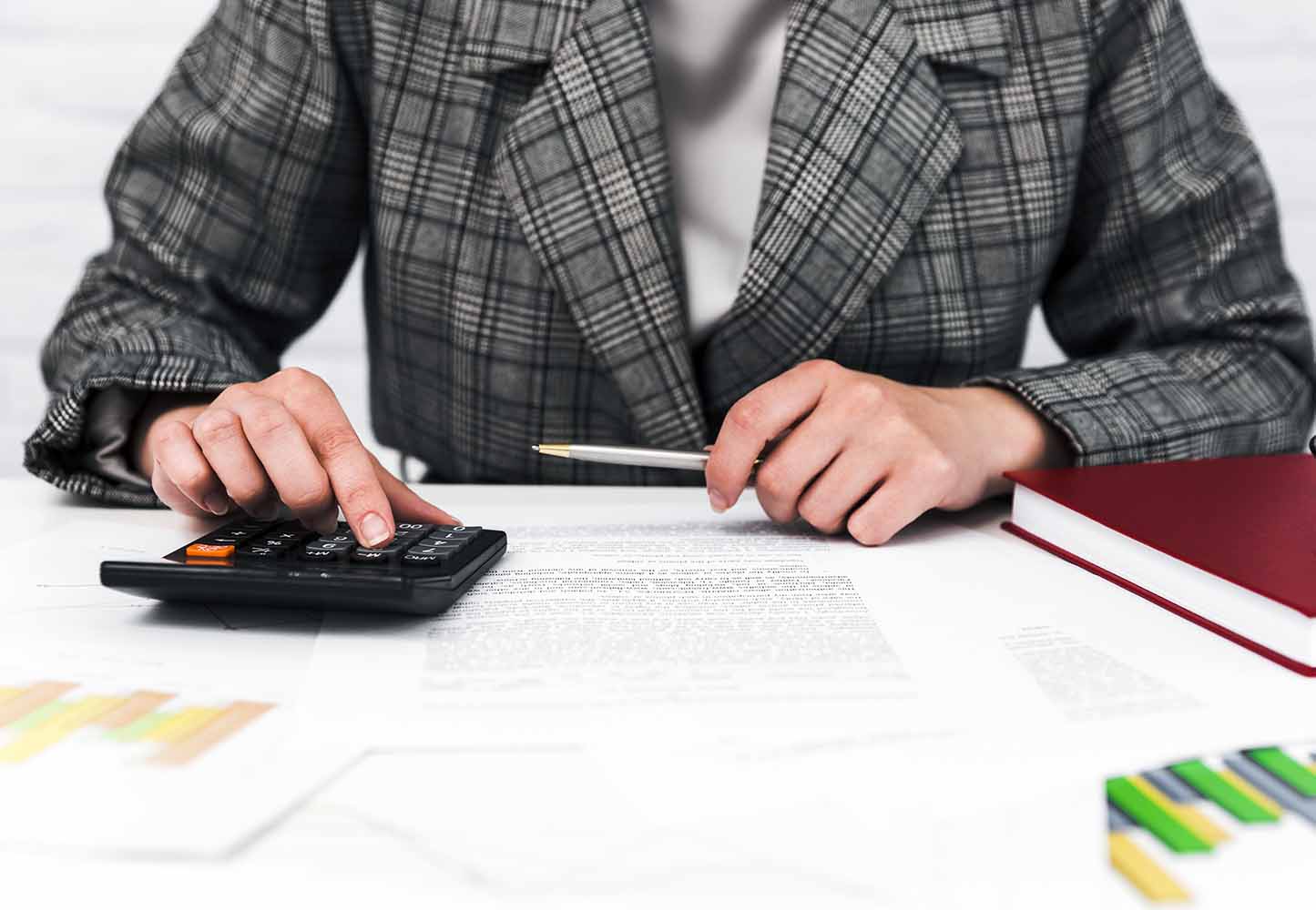 Person in a plaid blazer uses a calculator and writes on documents at a desk suggesting accounting work