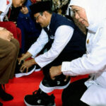 Volunteer in a white shirt helps a man put on black sneakers on a red carpet with others nearby