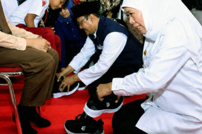 Volunteer in a white shirt helps a man put on black sneakers on a red carpet with others nearby