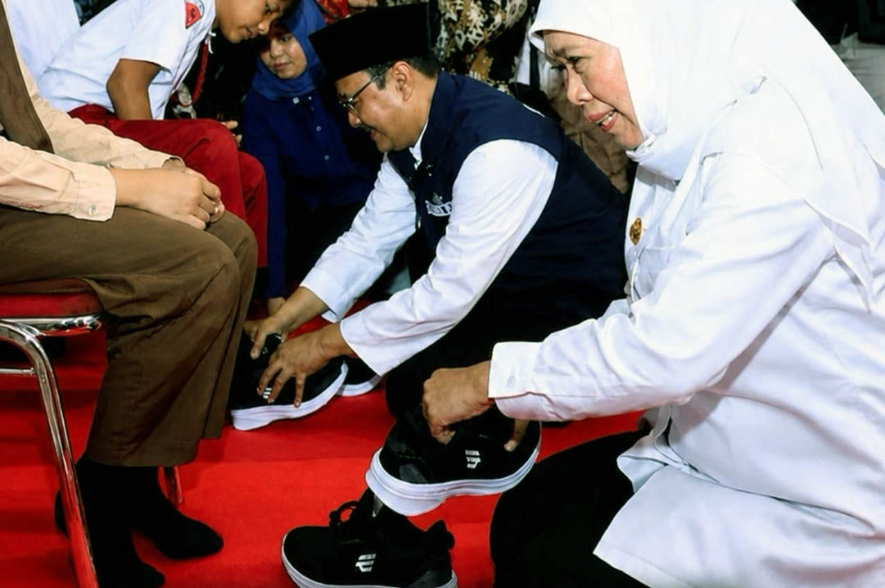 Volunteer in a white shirt helps a man put on black sneakers on a red carpet with others nearby