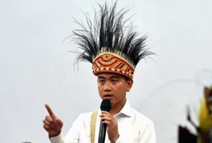 Man wearing a tall ornate feather headdress speaks into a handheld microphone gesturing with his index finger An event setting is visible in the background
