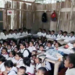 A classroom filled with students in white uniforms seated on the floor and at desks along the walls inside a bamboo walled room