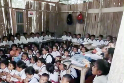A classroom filled with students in white uniforms seated on the floor and at desks along the walls inside a bamboo walled room