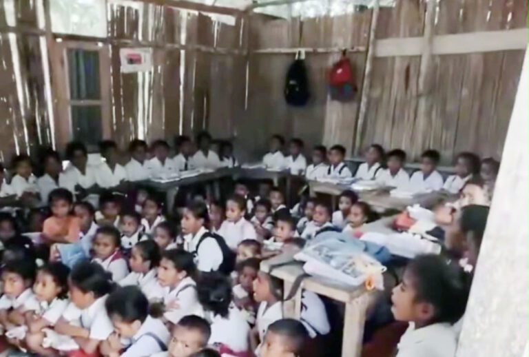 A classroom filled with students in white uniforms seated on the floor and at desks along the walls inside a bamboo-walled room.