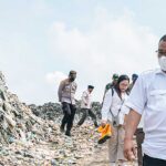People in masks and uniforms walk along a large garbage dump during a cleanup effort assessing waste piles nearby