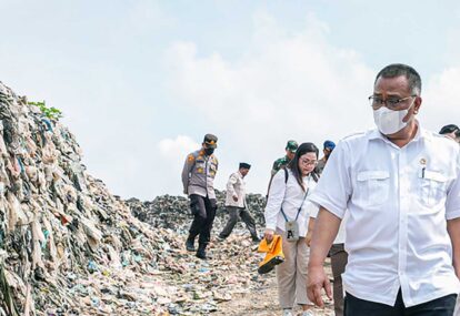 People in masks and uniforms walk along a large garbage dump during a cleanup effort assessing waste piles nearby