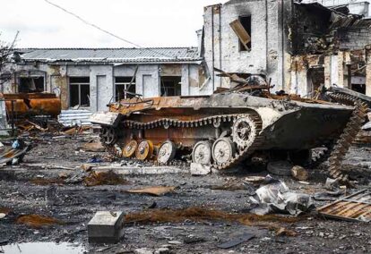 Rusted military tank wreck in a destroyed urban street with rubble and collapsed buildings behind it