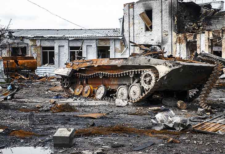 Rusted military tank wreck in a destroyed urban street with rubble and collapsed buildings behind it.