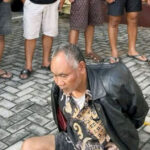 Man kneeling on a brick paved surface with his hands behind his back while several people stand around him in casual shoes and shorts
