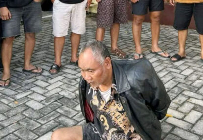 Man kneeling on a brick-paved surface with his hands behind his back, while several people stand around him in casual shoes and shorts.